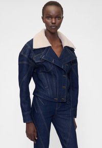 Tume denimist jakk pehme beežika kraega, topelt nööpidega, dekoratiivse õmblusega ja vormitud õmblustega. Liibuv lõige lühendatud disainiga.