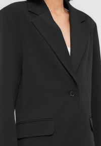 Γυναίκα με μαύρο, κομψό σακάκι με ένα κουμπί και πλαϊνές τσέπες, πάνω από λευκό εσωτερικό ρούχο, εμφανίζεται από τους ώμους έως τη μέση.