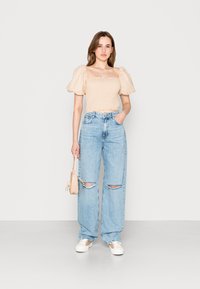 Haut court beige à manches bouffantes avec un design plissé, associé à un jean déchiré taille haute bleu clair et des baskets blanches. Tenant un petit sac beige.