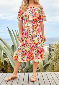 Robe fleurie décolletée avec une taille cintrée et un ourlet volanté, présentant des motifs vibrants rose, jaune et bleu. Portée avec des sandales plates.