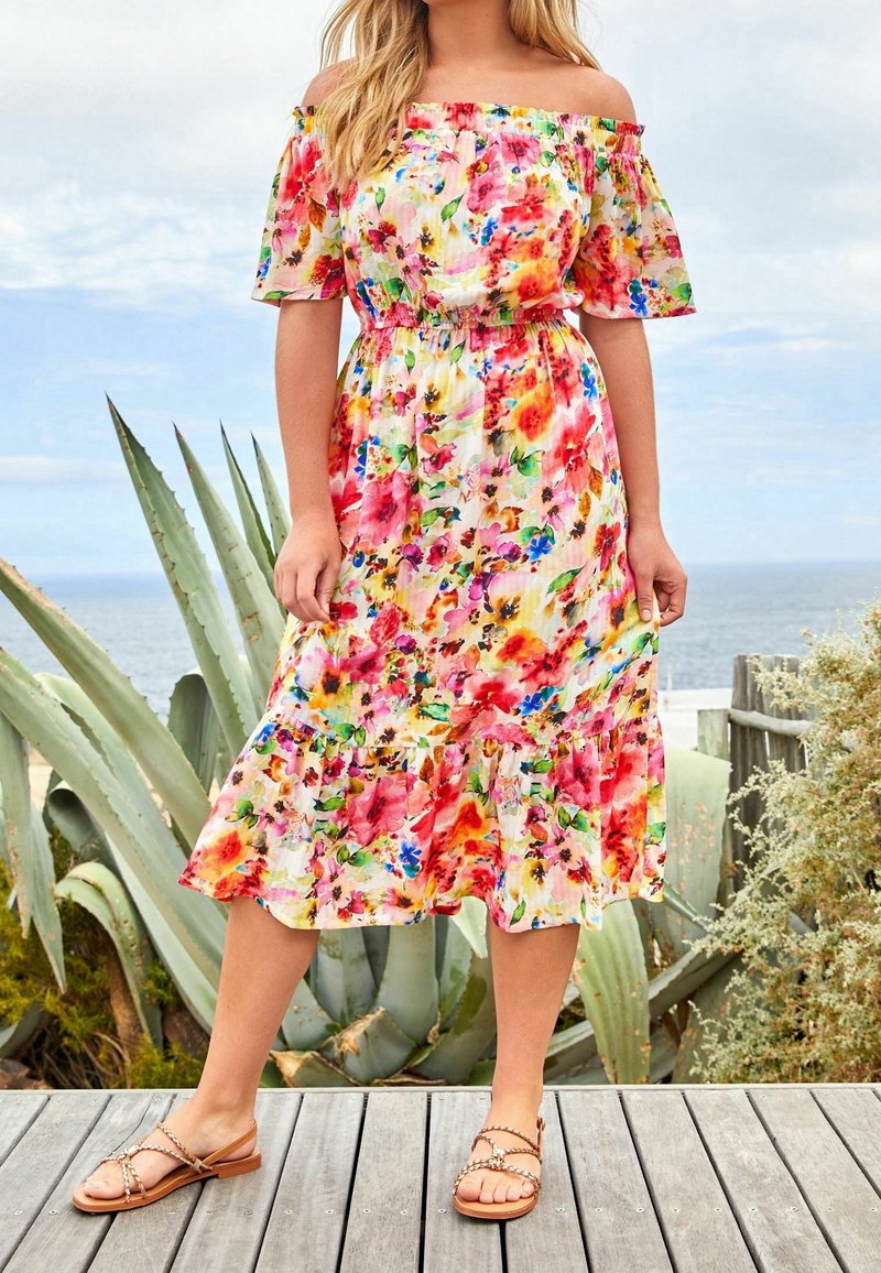 Robe fleurie décolletée avec une taille cintrée et un ourlet volanté, présentant des motifs vibrants rose, jaune et bleu. Portée avec des sandales plates.