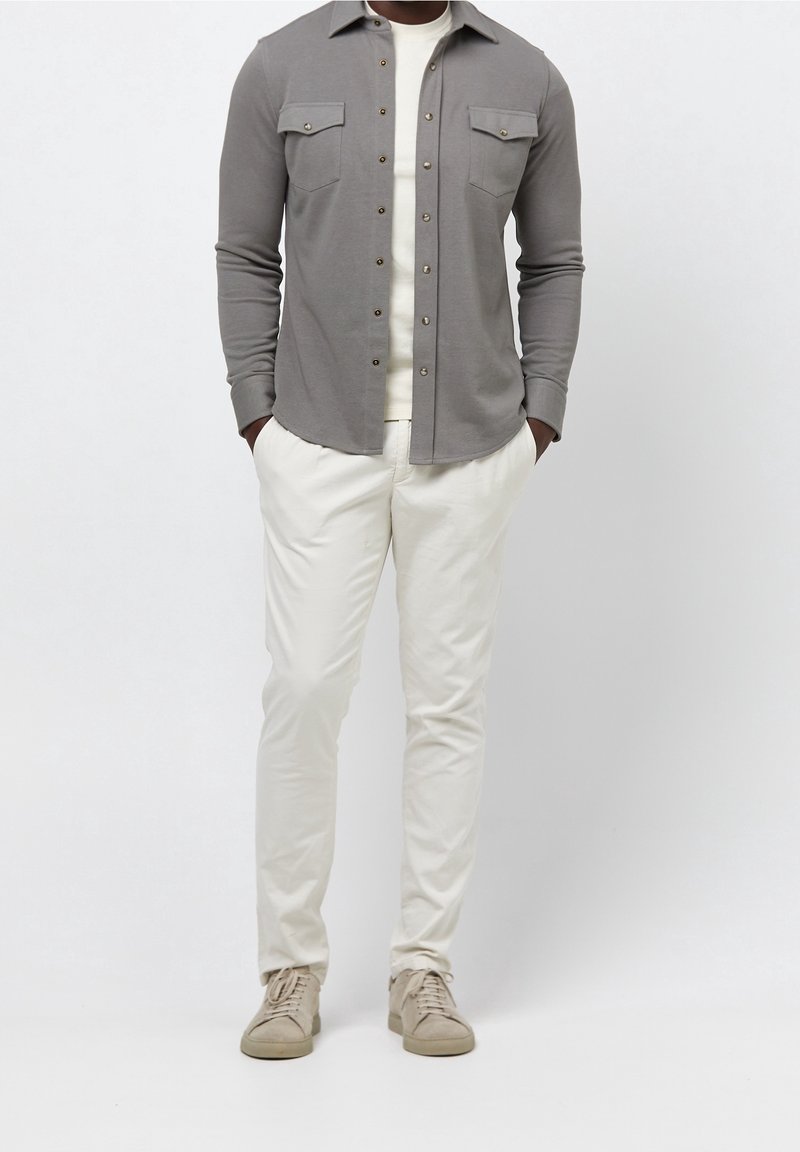 Chemise grise à manches longues avec col boutonné et deux poches poitrine, associée à un pantalon blanc et des baskets beige clair. Texture lisse.