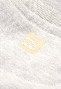Gros plan d'un tissu gris clair avec une étiquette carrée beige cousue portant l'inscription « NEW STEPS A NEW JOURNEY J&JOY ».
