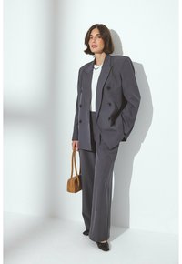 Completo grigio su misura con giacca doppiopetto, pantaloni a gamba ampia, top bianco e scarpe scure. Tiene una piccola borsa marrone. Texture liscia.