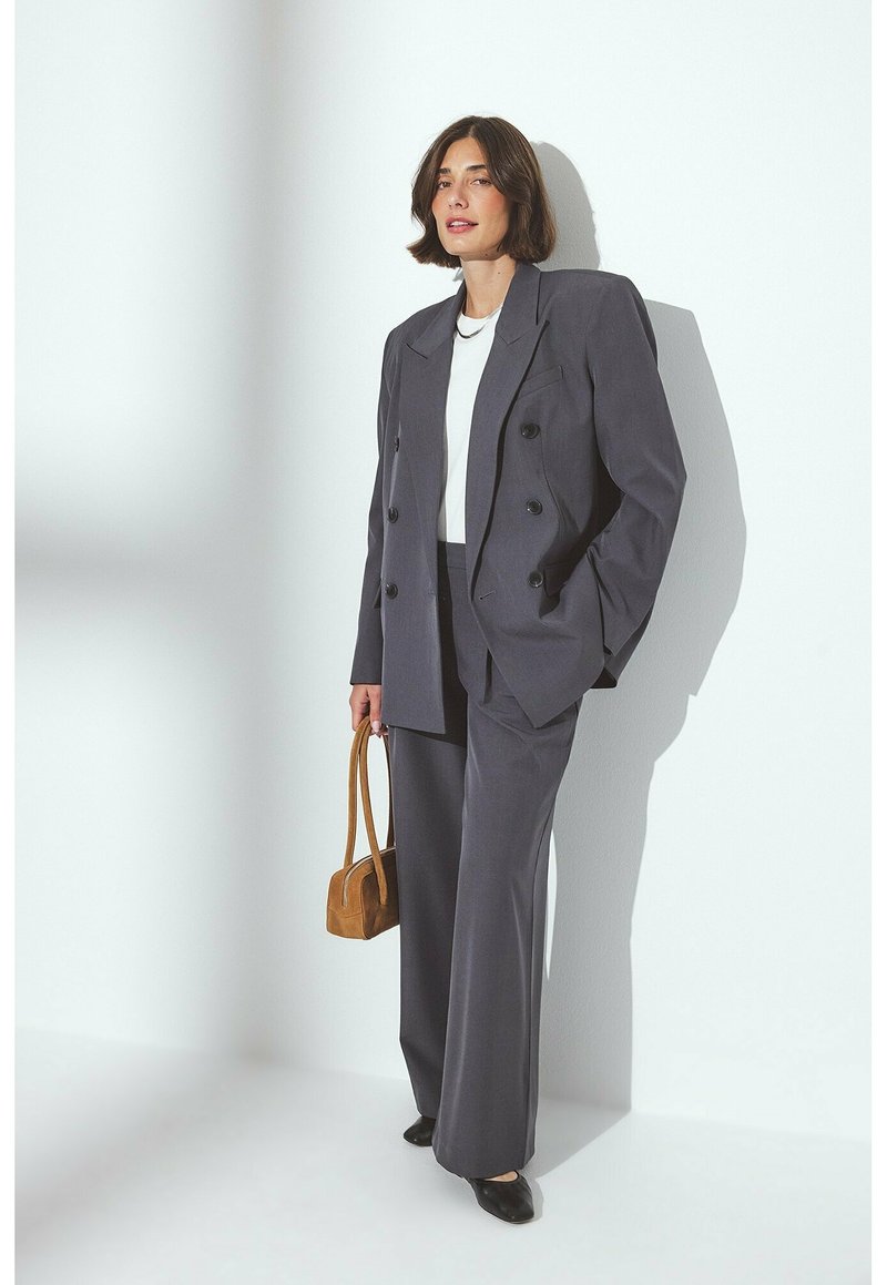 Completo grigio su misura con giacca doppiopetto, pantaloni a gamba ampia, top bianco e scarpe scure. Tiene una piccola borsa marrone. Texture liscia.