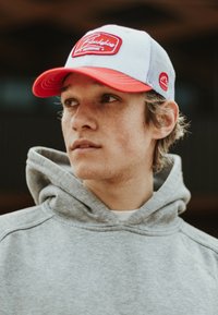 Valkoinen ja punainen baseball-lippis, jossa on verkkotakki ja logopatchi. Harmaa huppari näkyvissä, kangas näyttää pehmeältä, rento malli ja suuri huppu.