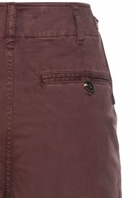 Gros plan sur un pantalon marron foncé montrant des passants de ceinture cousus et une poche arrière boutonnée sur une surface en tissu lisse.