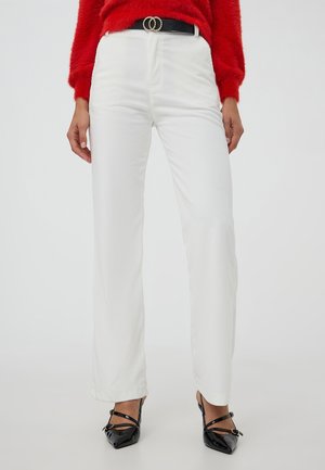 Donna che indossa pantaloni bianchi in corda a vita alta, una cintura nera con doppio anello, un maglione rosso peloso e tacchi a spillo neri con lacci su uno sfondo bianco.