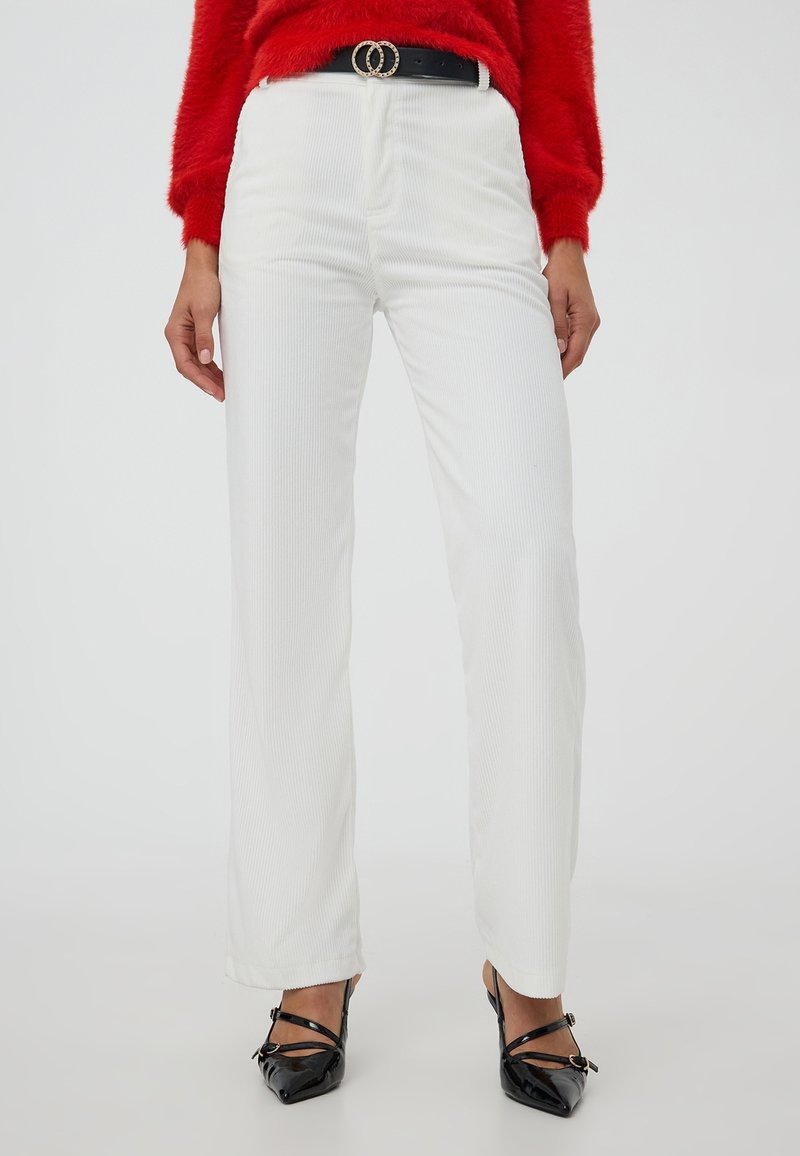 Donna che indossa pantaloni bianchi in corda a vita alta, una cintura nera con doppio anello, un maglione rosso peloso e tacchi a spillo neri con lacci su uno sfondo bianco.