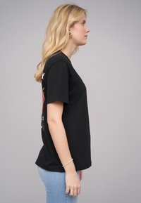 T-shirt noir en coton à manches courtes, avec un imprimé graphique dans le dos. Une paire de jeans bleu clair visible à la taille.