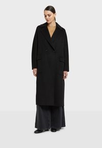 Čierny dvojradový vlnený kabát s klopovým golierom, dvoma prednými vreckami a nadrozmerným strihom. Nosený cez béžový top a čierne široké nohavice.