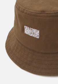 Brun bøttehatt laget av stoff med vertikal ribbing. Har hvit brodert merkenavn på forsiden og en bred, flat skygge.