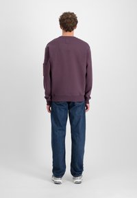 Ljubičasti dukserica s okruglim izrezom i dugim rukavima, koja ima prednji džep na lijevom rukavu. U kombinaciji s širokim plavim trapericama i tenisicama.