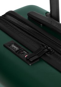 Valigia rigida verde con superficie testurizzata, doppie cerniere nere, serratura combinata integrata e manico retrattile.