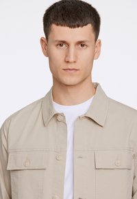 Ljusbeige jacka i tyg; har en spetsig krage, två framfickor och knappstängning. Bärs över en vit t-shirt.