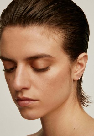 Un primer plano de la cara de una persona con pelo corto y mojado de color castaño; piel suave; maquillaje natural; y un pequeño pendiente brillante en la oreja derecha.