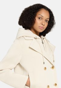 Beiger doppelreihiger Mantel mit übergroßem Kapuzenet, verziert mit goldfarbenen Knöpfen und glatter Textur. Verfügt über Seitentaschen und Kordelzugdetails.