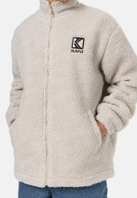 Krämfärgad fleecejacka med en strukturerad, mjuk yta. Har en ståkrage, dragkedjestängning och en broderad logotyp på bröstet.