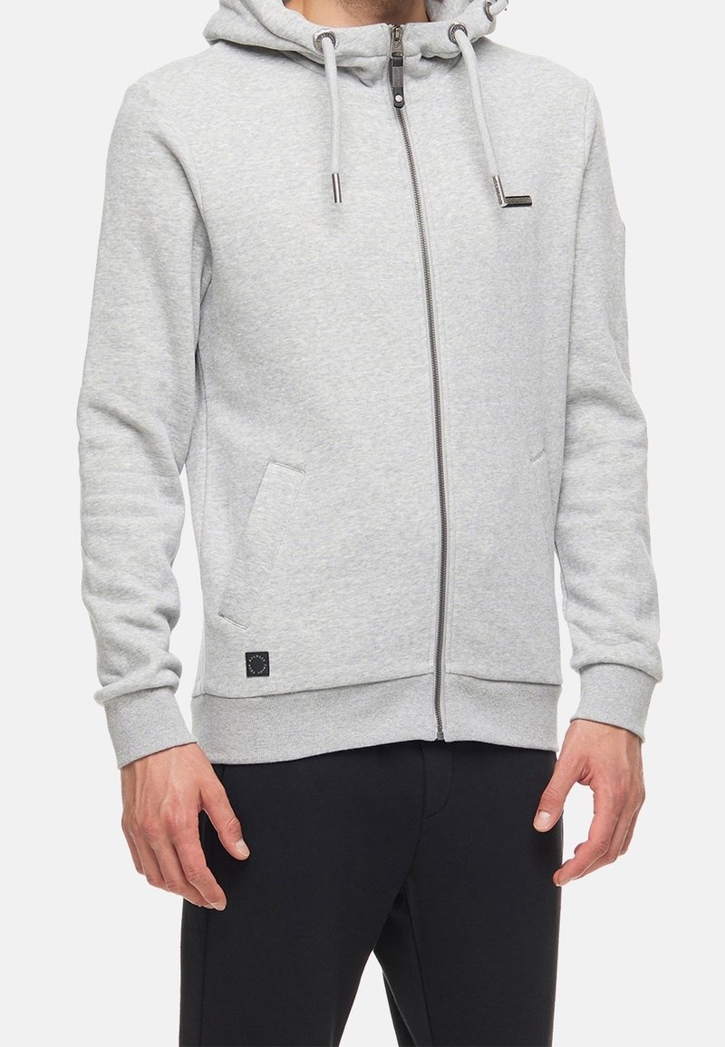Personne portant un sweat à capuche gris clair zippé avec cordons et pantalon noir, debout les bras détendus sur les côtés.