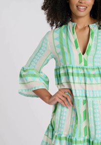 El vestido estampado presenta rayas geométricas en verde claro y turquesa, con un escote en V y mangas acampanadas, hecho de una tela ligera.