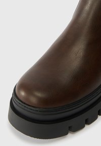 Brun läderankelboot med rund tå, texturerad yta och en kraftig svart gummisula med horisontella spår.