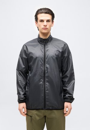 Noormees näoga ettepoole, kandes musta kerget lukuga jope ja oliivrohelisi cargo-pükse, taustaks ühtlane ilma.