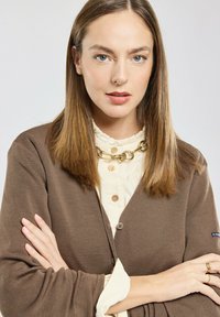 Cardigan marrone a coste con scollo a V, bottoni frontali e maniche lunghe, indossato sopra una blusa color crema con colletto ondulato e collana d'oro.