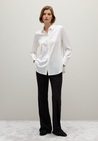 Camisa blanca de cuello con botones y mangas largas, combinada con pantalones negros de pierna ancha y zapatos negros decorados sobre una superficie de color claro.