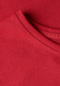 Tissu en coton rouge superposé avec une texture lisse. Il présente un col rond et des bords droits, avec des détails de couture visibles.