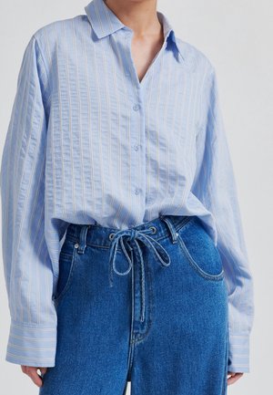 Person iført en lyseblå stripet knap-op skjorte stoppet ned i højttaljede blå jeans med et bundet denim bælte.