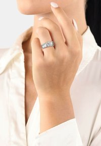 Anello in argento con tre file di bande testurizzate, caratterizzato da un mazzo centrale di pietre chiare. Finitura liscia, indossato su una mano con una leggera manicure.