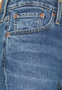 Gros plan d'une poche de jean en denim bleu avec rivets en cuivre et coutures jaunes sur un tissu texturé.