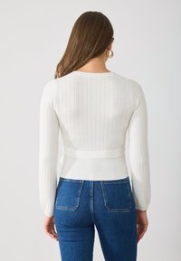 Top lung cu maneci lungi, alb cu un elastic strâmt în talie, asortat cu blugi din denim albastri cu buzunare pe spate și cusături standard.