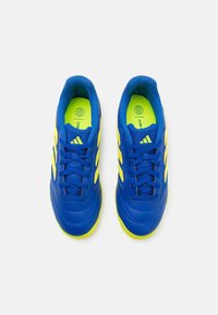 Ett par blåa sportskor med gula ränder och neongula sulor, vy från ovan på en vit bakgrund.