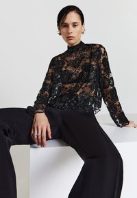 Schwarzes Spitzenoberteil mit Blumenmustern, hohem Kragen und langen Ärmeln; kombiniert mit schwarzen, hoch taillierten Hosen mit glatter Textur.