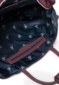 Tummanpunaisen laukun sisäosa, jossa on tummansininen vuori, toistuva valkoinen U.S. Polo Assn. -logokuvio, vetoketjullinen tasku ja sileä pinta.