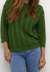 Grüner, kabelgewickelter Pullover mit 3/4-Ärmeln, der über strukturierte Muster und einen runden Halsausschnitt verfügt, kombiniert mit hellblauen Denim-Jeans.