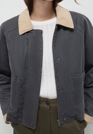 Femme portant une veste gris foncé avec un col et des poignets en velours côtelé beige, un haut en maille blanc et un pantalon taille haute vert olive.