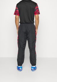 Svart sportoutfit bestående av en T-shirt med röda geometriska mönster och matchande svarta byxor, detaljerade med röda accenter och sidostråk. Blå skor.