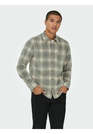 Camicia a quadri verdi con maniche lunghe, dotata di tasca sul petto e bottoni sul davanti, indossata con pantaloni scuri.