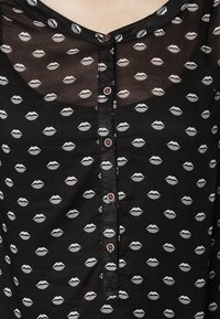 Blouse noire en tissu transparent avec un motif répétitif d'imprimé lèvres. Présente un devant boutonné avec des boutons rouges. Tissu doux et léger.