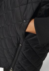 Hand in de zak van een zwarte gewatteerde jas met drukknoop en rits, waarbij de mouwzoom van gestructureerde zwarte stof zichtbaar is.