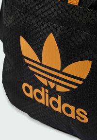 Czarna siatkowa torba o fakturze plastra miodu, z pomarańczowym logo w kształcie trzech liści i wyraźnie widoczną nazwą marki z przodu.