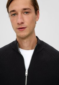 Schwarzer gerippter Pullover mit einem halben Reißverschlusskragen und silbernem Reißverschluss. Über einem weißen Hemd getragen, mit einer glatten Textur und taillierter Passform.