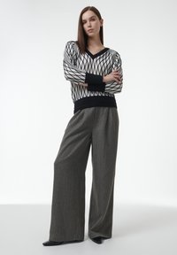 Schwarz-weißer, rautenförmiger Pullover mit V-Ausschnitt, kombiniert mit grauen, hoch taillierten, weiten Hosen und schwarzen spitzen Absätzen.