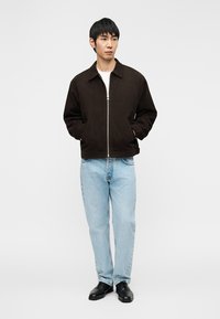 Καφέ σουέντ μπουφάν μπέρτζερ με ασημένιο φερμουάρ, φορεμένο πάνω από λευκό πουκάμισο, σε συνδυασμό με ανοιχτό μπλε τζιν και μαύρα παπούτσια.