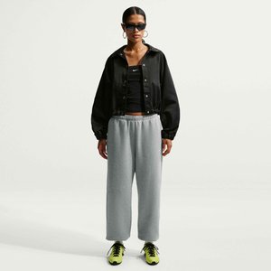 Vrouw met een zwart cropped jack, zwart cropped top, grijze wijde sweatpants, neongele sneakers, zonnebril en oorringen, staand op een witte achtergrond.