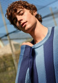 Blauer und Marine gestreifter Strickpullover aus strukturiertem Material, mit geripptem Kragen und Bündchendetails. Lässige Passform, die schulterfrei gegen einen Zaun getragen wird.
