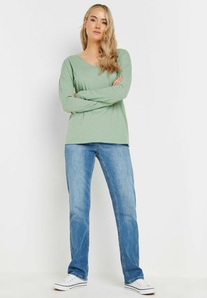 Langærmet, v-hals top i lys grøn, parret med blå jeans og hvide sneakers. Stoffet ser letvægts ud med en blød tekstur.