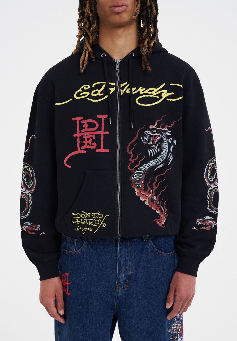 Felpa con zip nera caratterizzata da grafiche dettagliate di un drago e serpenti. Il materiale sembra morbido. Include loghi con accenti gialli e rossi.